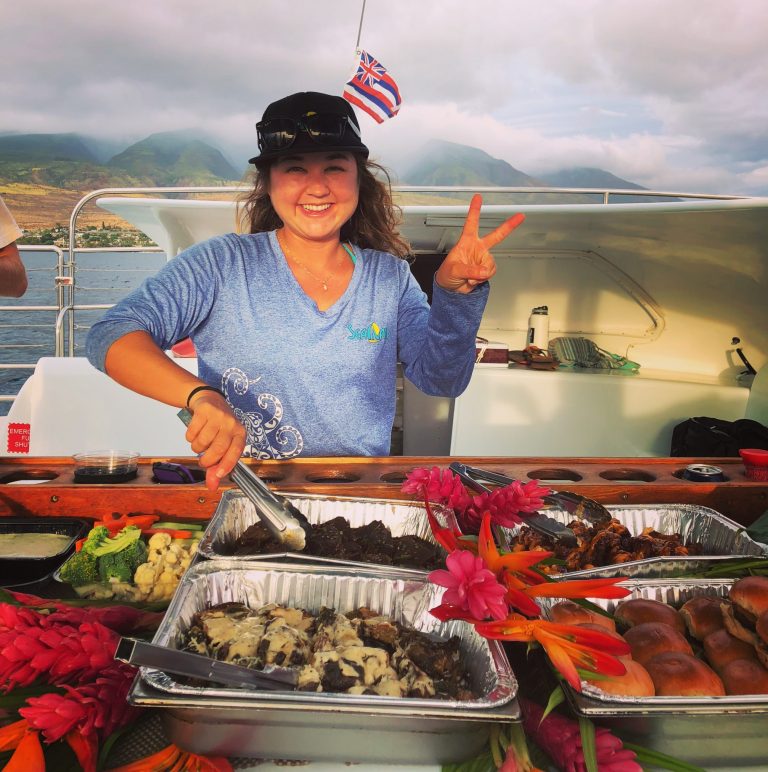 Smiling person on a boat serving food with a peace sign, colorful flowers and mountains in the background.