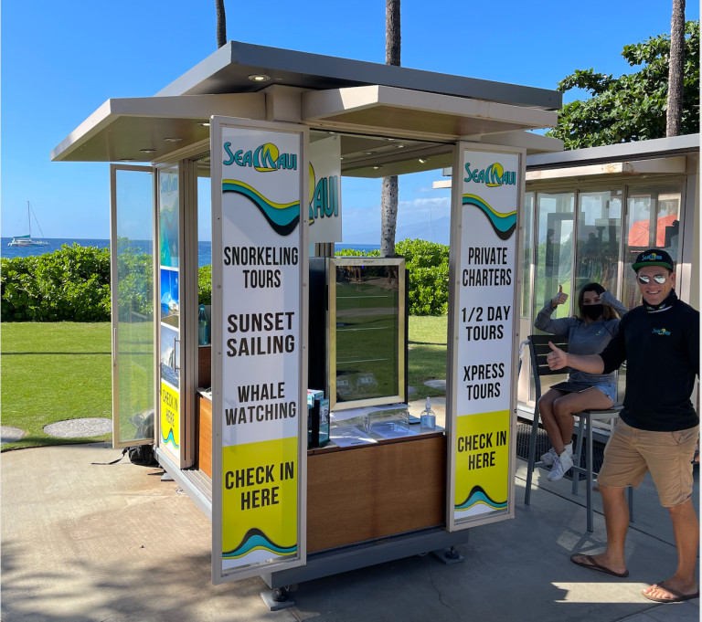People standing near a tour booking kiosk by the beach with signs for various activities.