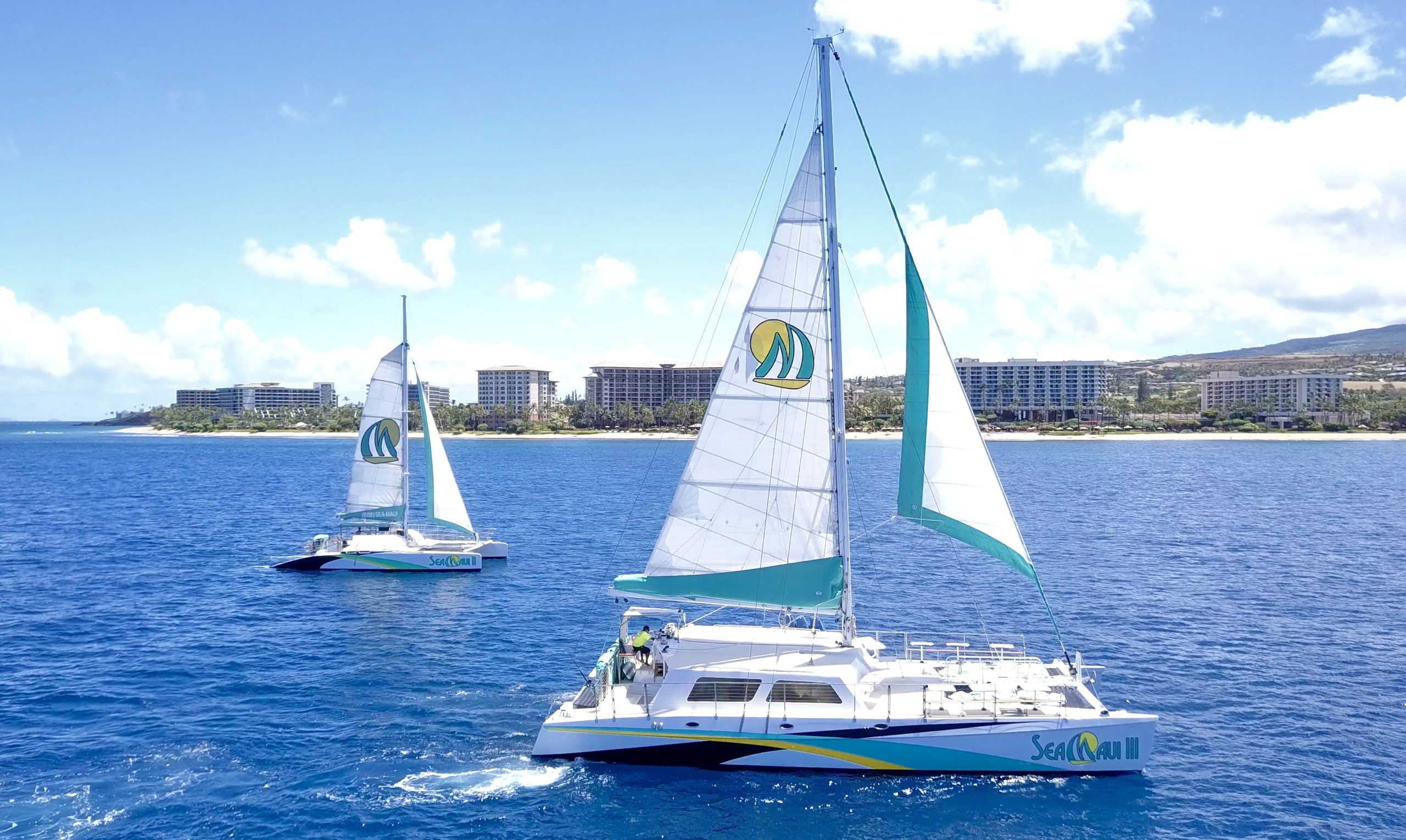 Two sailboats on blue sea with hotels and coastline in the background.