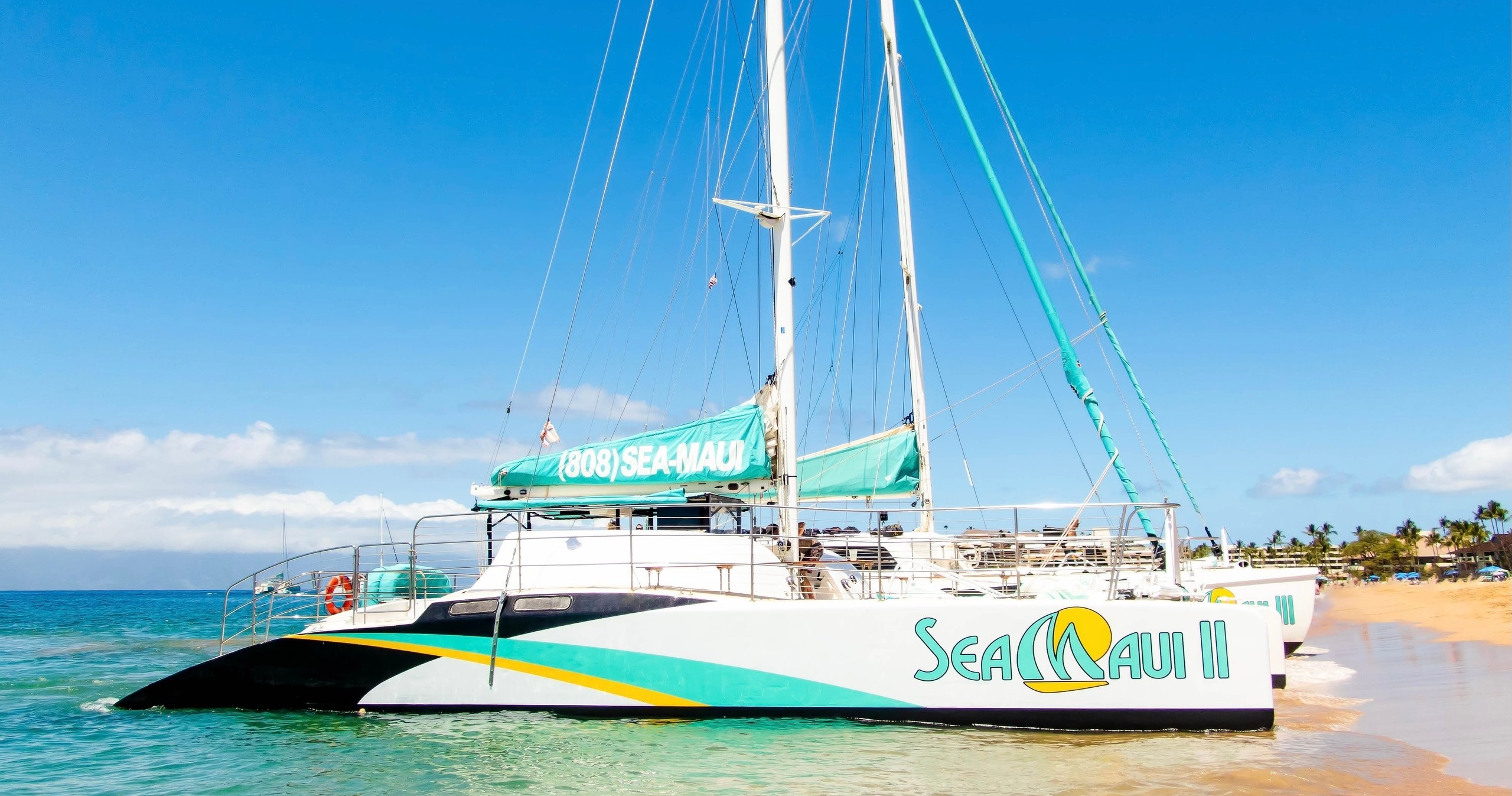 Catamaran named 'Sea Maui II' on beach with blue sky.