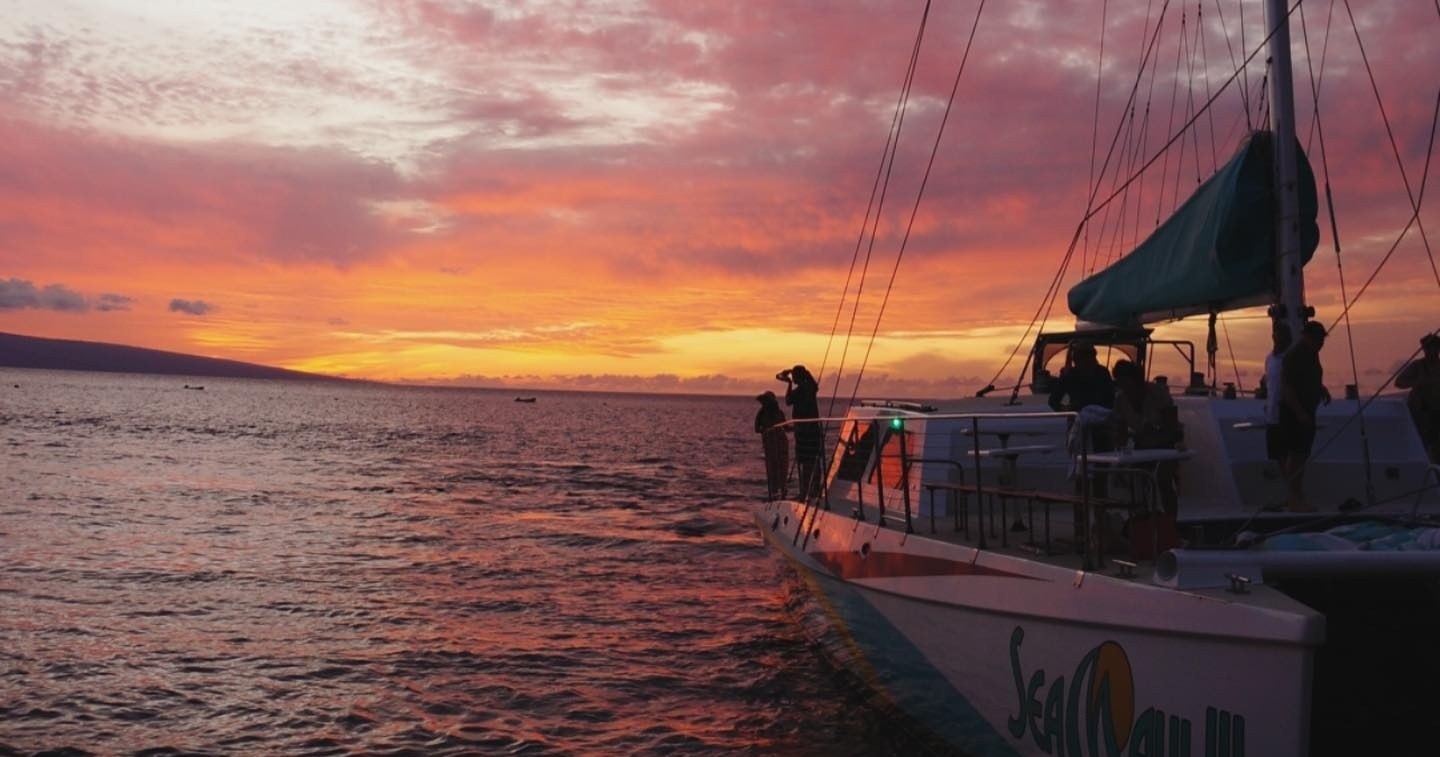Boat with people on water at sunset, sky in vibrant orange and pink hues.