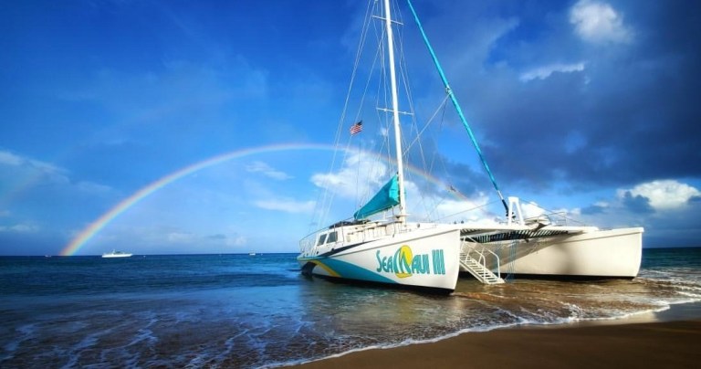 Catamaran on beach with rainbow over ocean under blue sky.
