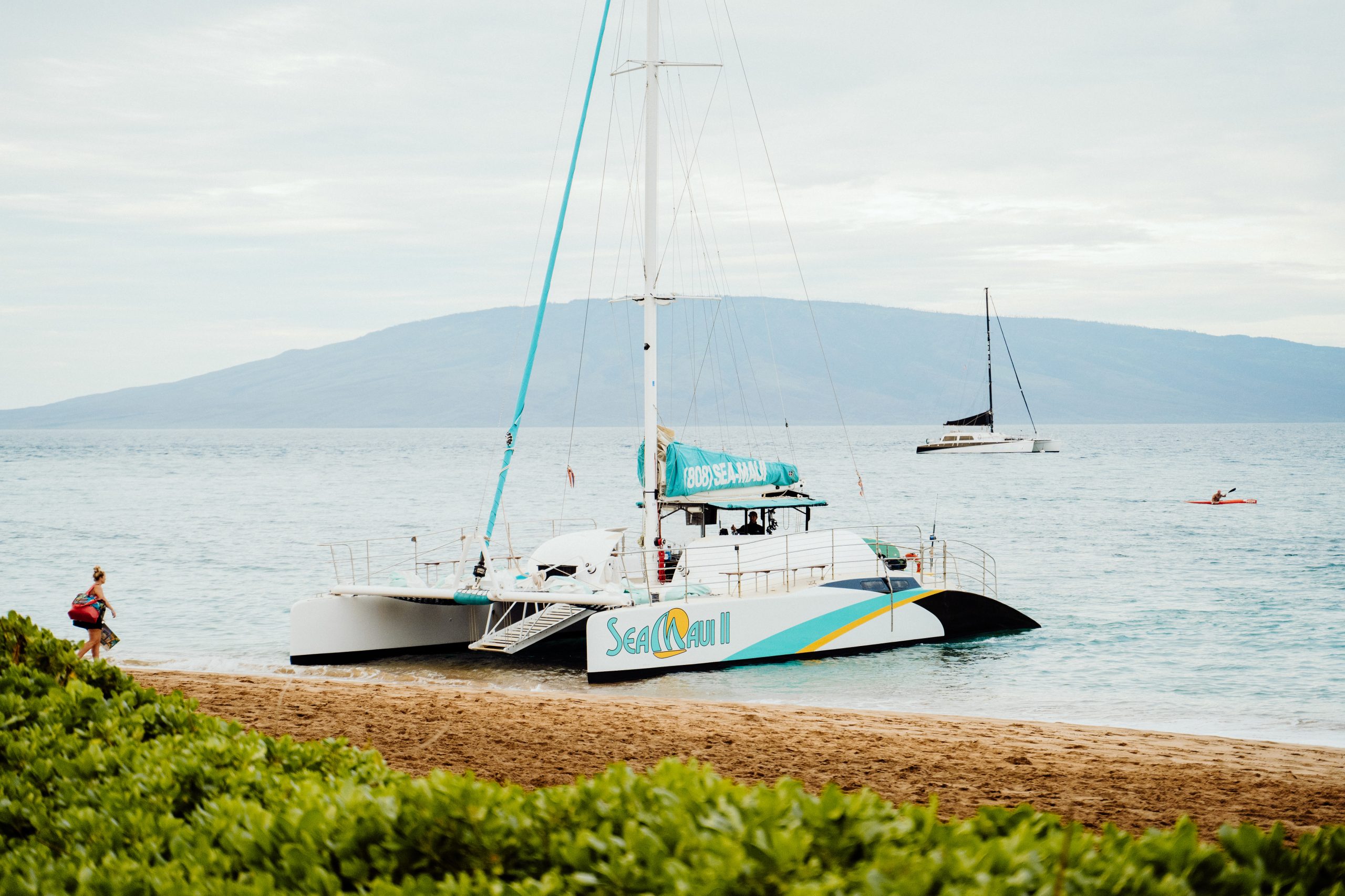 Catamaran on beach with mountains and sailboat in background.
