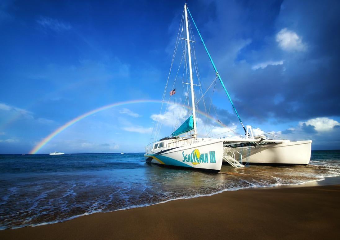 Catamaran on beach with rainbow in a cloudy sky.