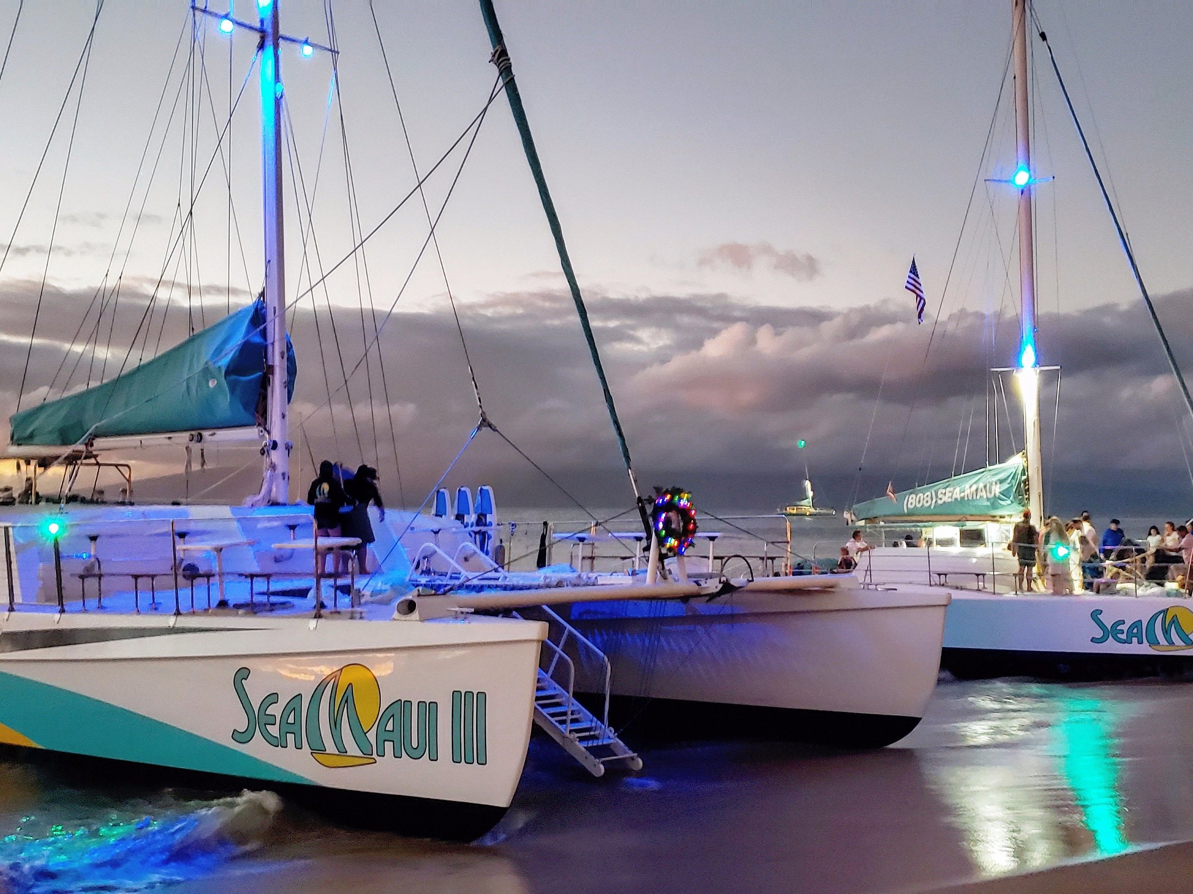 Two catamarans docked on a beach at dusk with lights on mast, clouds in background.