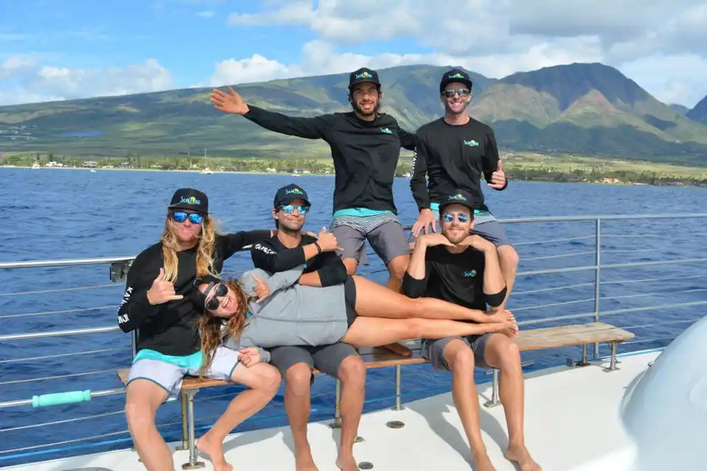 Group of six people in matching outfits posing on a boat with mountains in the background.