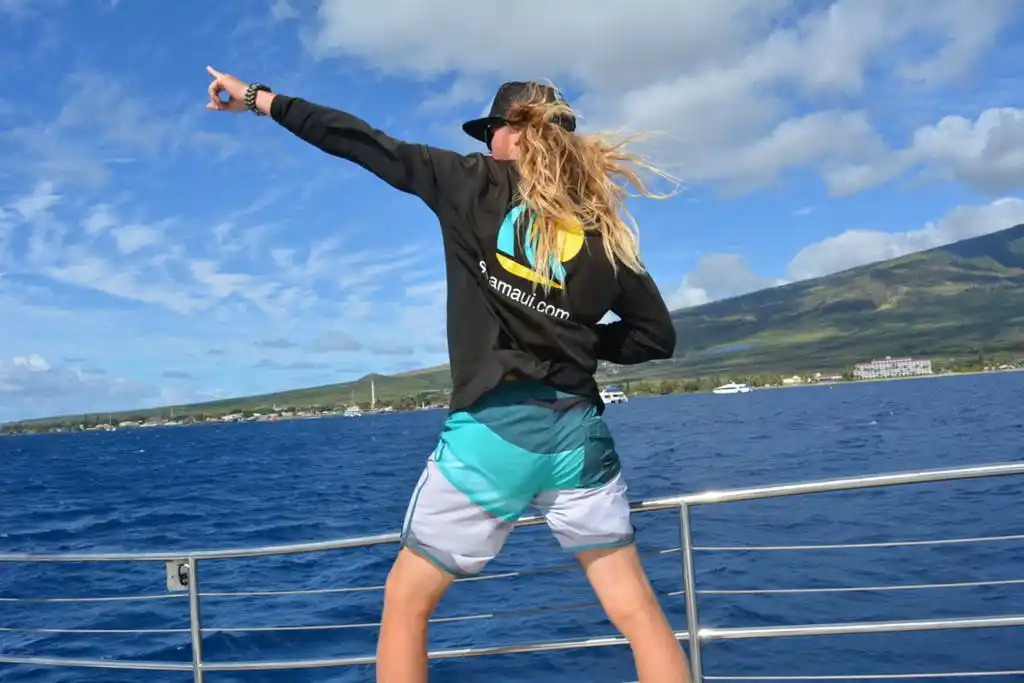 Person with long hair pointing towards sky on boat, wearing hat and jacket.