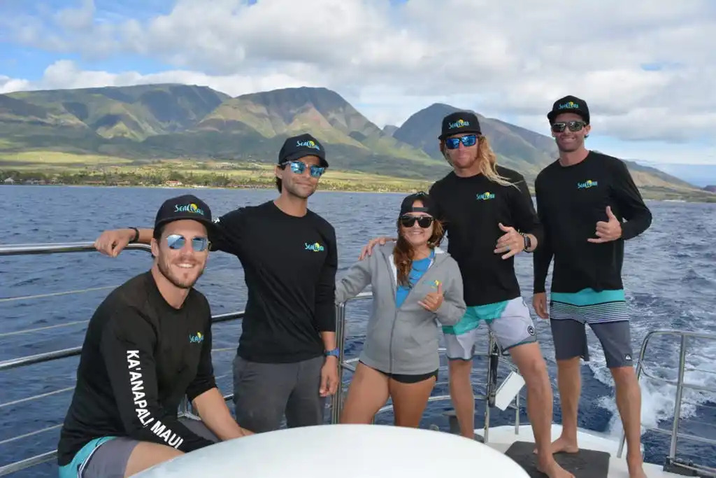 Group of five people on a boat, wearing sunglasses and caps, with mountains in the background.