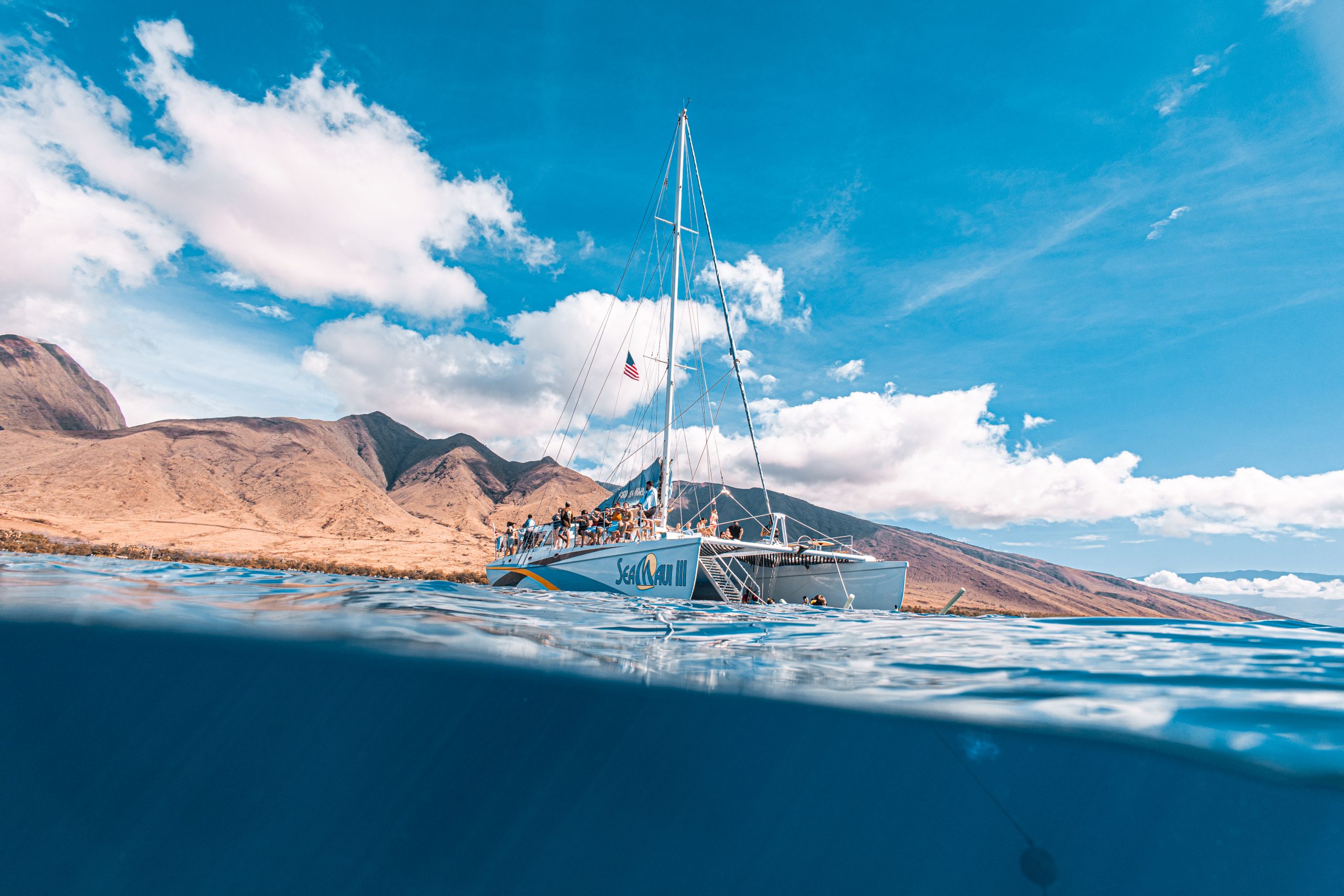 Catamaran with people sails near mountains under blue sky.