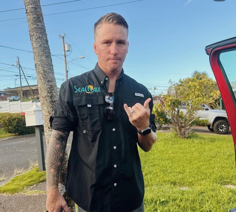 Man with tattoos in black shirt making a hand gesture near a car, outdoors.