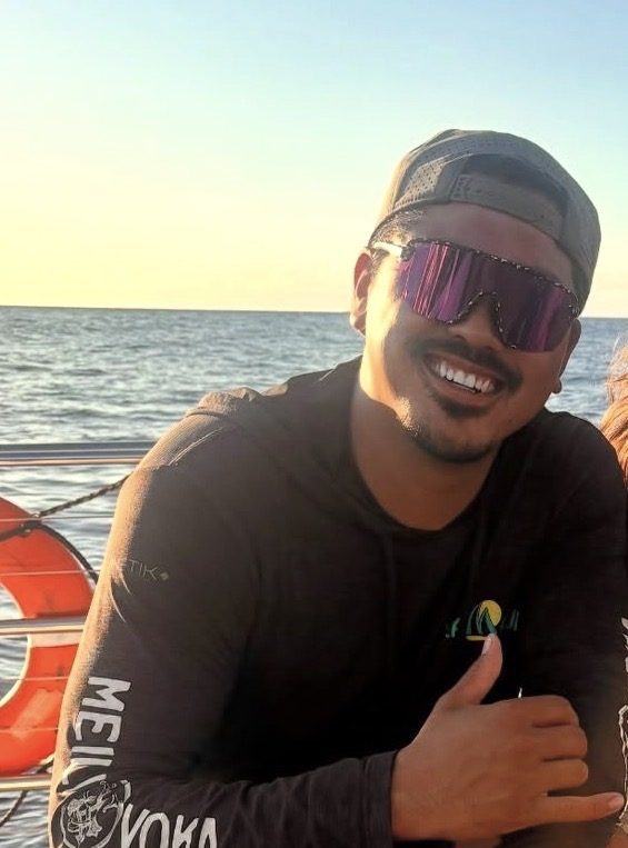 Man on a boat wearing sunglasses and a cap, holding a drink, smiling at sunset.