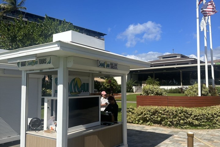 Small outdoor kiosk on sidewalk near bushes, with person inside.