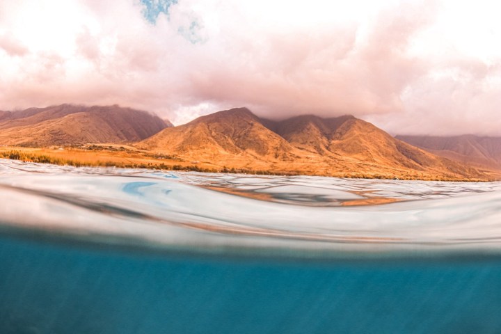 Brown mountains and cloudy sky above blue ocean with surface visible.