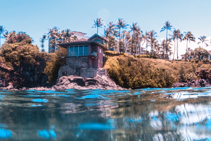 House on a cliff with palm trees, viewed from waterline.