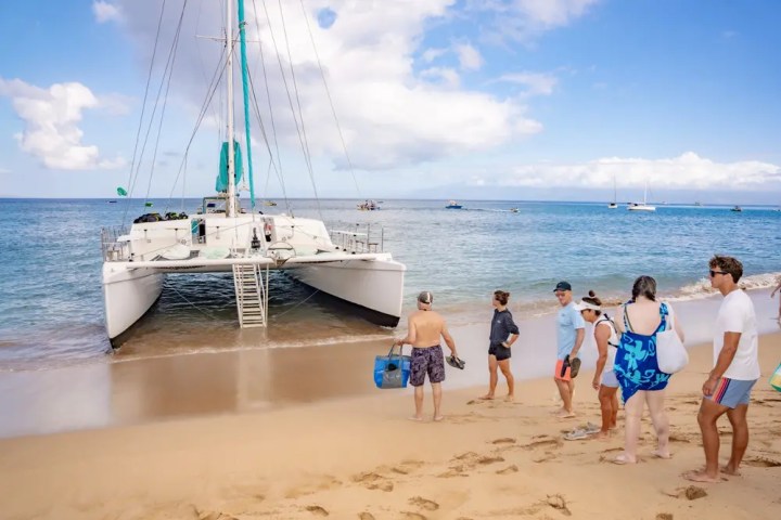 People boarding a catamaran on a sandy beach under a blue sky.